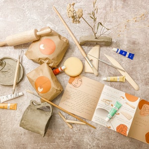 May include: A pottery tool kit with various supplies for clay modeling. Includes a wooden rolling pin, paint tubes, brushes, a sponge, and a booklet with instructions. The kit also features a small, gray clay vase and a round, gray clay dish.