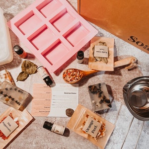 May include: A flat lay of soap-making supplies. Includes a pink silicone mold, clear soap bars with botanicals, essential oil bottles, a wooden spoon with dried fruit, and small paper bags of ingredients. A box with "Soak" printed on it is also visible.