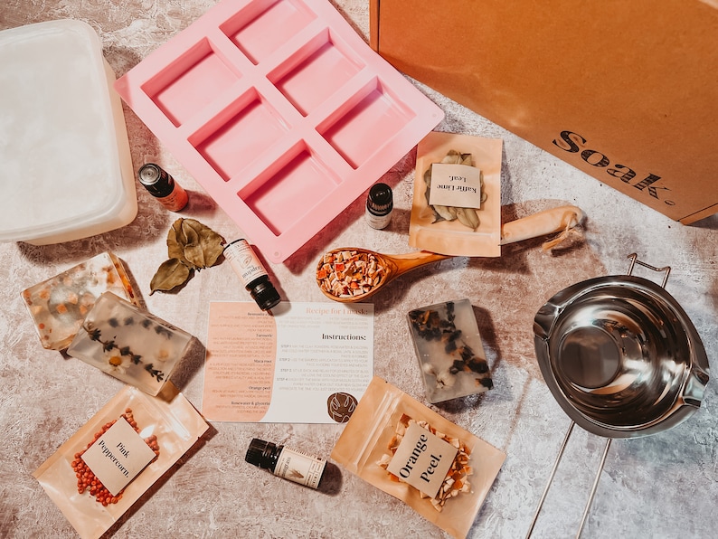 May include: A flat lay of soap-making supplies. Includes a pink silicone mold, clear soap bars with botanicals, essential oil bottles, a wooden spoon with dried fruit, and small paper bags of ingredients. A box with "Soak" printed on it is also visible.