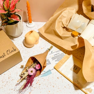 May include: A craft kit with Jesmonite powder and liquid, cups, and molds, alongside a small bouquet of dried flowers and a decorative candle. A box with "The Cast Club" is also visible. The kit is presented on a speckled surface.