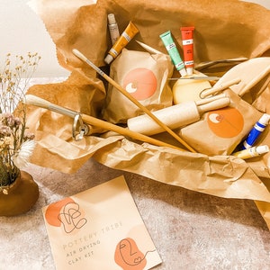 May include: A Pottery Tribe air-drying clay kit in a brown paper-lined box. The kit includes paint tubes, brushes, wooden tools, and clay. A small vase with dried flowers is on the left. The kit's packaging has a logo with an orange circle and abstract design.