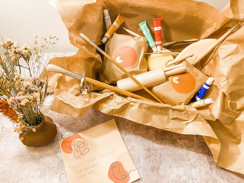 May include: A Pottery Tribe air-drying clay kit in a brown paper-lined box. The kit includes paint tubes, brushes, wooden tools, and clay. A small vase with dried flowers is on the left. The kit's packaging has a logo with an orange circle and abstract design.