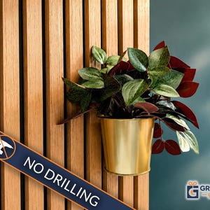 May include: A gold-colored metal plant pot mounted on a light brown vertical wooden slat wall. The pot contains a leafy plant with green and deep red foliage. A diagonal blue banner across the bottom displays the text NO DRILLING.