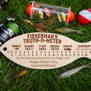 Könnte beinhalten: Ein fischförmiges Holzschild, "Fisherman's Truth-O-Meter", mit Angelködern, einer Rolle und einer Pose auf Gras. Das Schild hat eine Skala und den Text: "Happy Father's Day, Love from Richard."