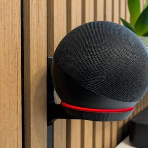 Pode incluir: Um alto-falante inteligente preto com um exterior de malha de tecido e uma base iluminada em vermelho. O alto-falante é montado em um suporte preto contra uma parede de ripas de madeira. Uma pequena planta em um vaso branco é visível ao fundo.