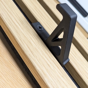 Può includere: Primo piano di una staffa in metallo nero fissata a doghe di legno di colore chiaro. La staffa è fissata con una singola vite e presenta un design angolare distintivo. Le doghe di legno sono disposte in parallelo.