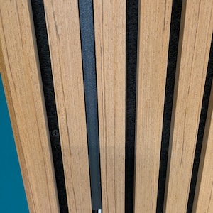 May include: A wall panel featuring vertical light brown wood slats separated by deep black felt gaps. A thin black cable management channel is mounted vertically between two slats, partially concealing a white power cord running through it.