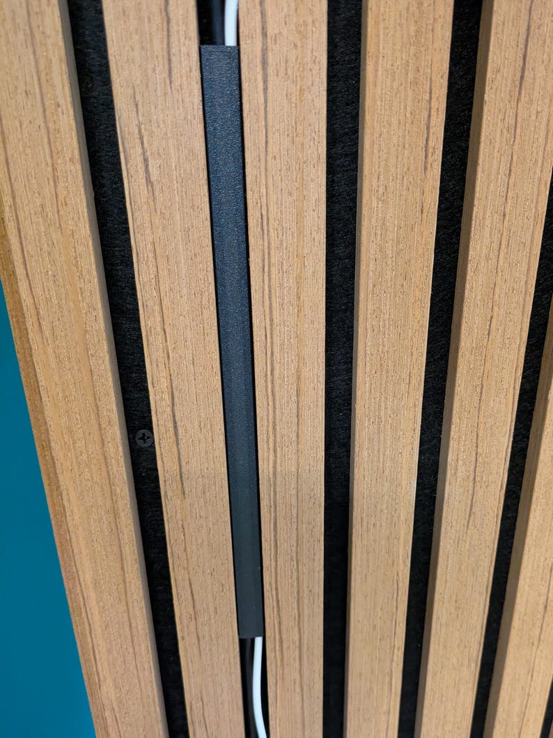 May include: A wall panel featuring vertical light brown wood slats separated by deep black felt gaps. A thin black cable management channel is mounted vertically between two slats, partially concealing a white power cord running through it.