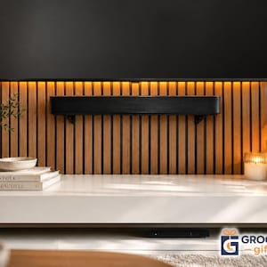 May include: A black soundbar mounted on a wall with vertical wood slats, positioned beneath a large flat-screen television with warm under-cabinet lighting. The setup includes two stacked books titled Kinfolk and Live Beautiful on a white shelf.