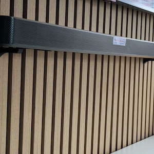 Puede incluir: Una barra de sonido larga y rectangular, de color gris oscuro, montada en una pared de listones de madera con soportes negros. La barra de sonido tiene un panel frontal perforado y una pequeña etiqueta. La pared es de color marrón claro con rayas verticales marrones oscuras.