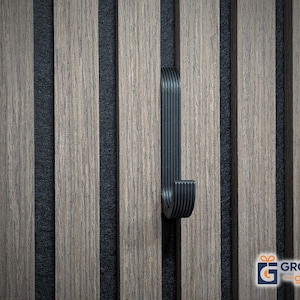 Puede incluir: Un gancho de metal negro está montado en un panel de madera vertical con tiras de fieltro gris oscuro alternas. El gancho tiene un diseño curvo con ranuras verticales. La veta de la madera es visible en los paneles de color marrón claro.