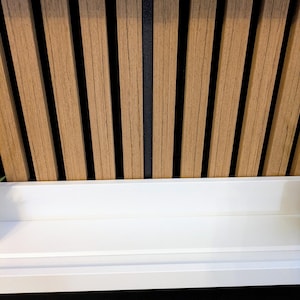 May include: A wall-mounted white shelf positioned in front of a vertical wood slat panel. The panel features light brown wooden slats separated by thin black gaps. Below the shelf, a black horizontal soundbar is visible with a small blue light.