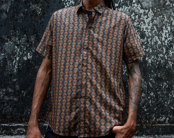 Op de afbeelding: Een man draagt een zwart shirt met oranje patroon, een zwarte pet en een zonnebril. Hij staat voor een getextureerde grijze muur.