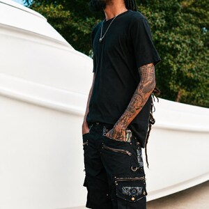 May include: A black t-shirt and black cargo shorts featuring multiple pockets with metallic zippers, decorative metal rings, and patterned fabric accents on the pocket flaps. The shorts have a structured design with visible stitching and hardware details.