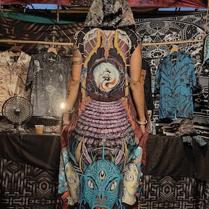 Op de afbeelding: Een lange, mouwloze, gekapte jurk met een kleurrijke, psychedelische print. De print bevat een groot, blauw, drieeogig demonen gezicht met een lange, oranje tong. De jurk is gemaakt van een zachte, comfortabele stof en heeft een ontspannen pasvorm.