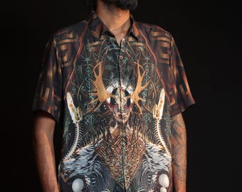 May include: Short-sleeved button-down shirt with a detailed, colorful graphic print. The design features a central figure with antlers, surrounded by geometric and organic patterns in shades of brown, blue, and white. The shirt has a classic collar and button closure.