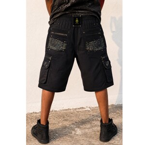 May include: Black cargo shorts featuring multiple pockets with silver-toned zippers and metallic studs. The back pockets display a detailed grey geometric pattern. A small rectangular black patch with a gold emblem is centered on the waistband.