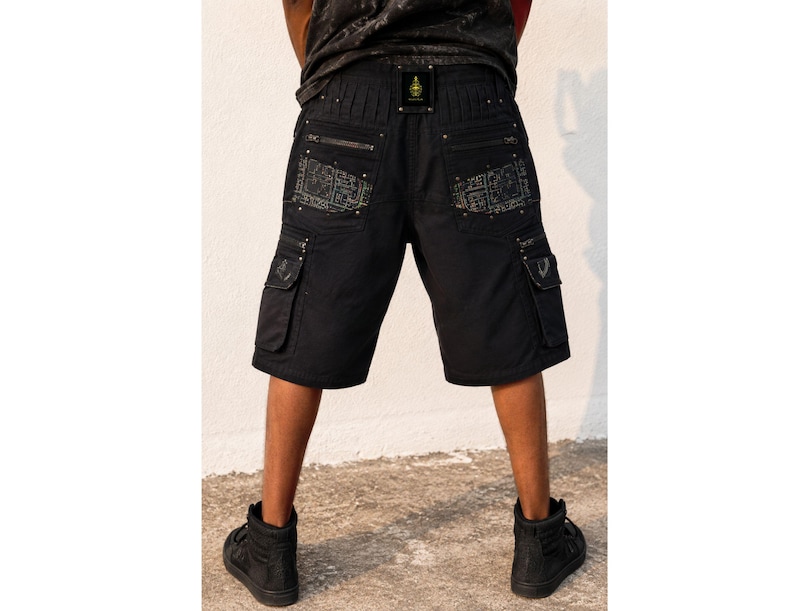 May include: Black cargo shorts featuring multiple pockets with silver-toned zippers and metallic studs. The back pockets display a detailed grey geometric pattern. A small rectangular black patch with a gold emblem is centered on the waistband.