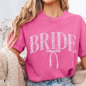 May include: A pink t-shirt with the word "BRIDE" in white script with a white bow underneath. The shirt is being worn by a person with long blonde hair.