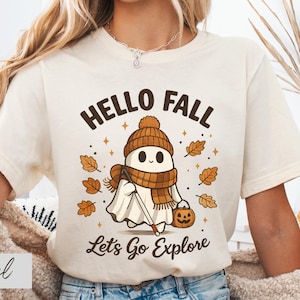 May include: A natural-colored t-shirt featuring a graphic of a ghost wearing a brown knit hat and scarf, holding a walking stick and a jack-o'-lantern bucket. The text reads HELLO FALL and Let's Go Explore, surrounded by falling autumn leaves.