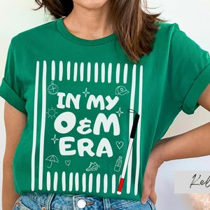 May include: A green t-shirt with the text "IN MY O&M ERA" in white, along with various small white illustrations. The shirt has rolled-up sleeves and is worn by a person.