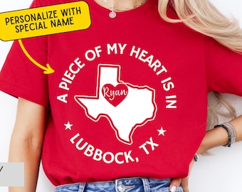 May include: Red t-shirt with a white graphic of the Texas state outline and a heart. The text reads "A PIECE OF MY HEART IS IN LUBBOCK, TX" with the name "Ryan" inside the heart. The shirt also has a yellow banner that says "PERSONALIZE WITH SPECIAL NAME".