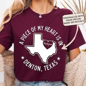 May include: A maroon t-shirt featuring a white graphic of the state of Texas with a heart icon containing the name Madyson. The text reads A PIECE OF MY HEART IS IN DENTON, TEXAS. A callout box says PERSONALIZE WITH YOUR SPECIAL NAME OR INITIALS.