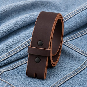 May include: A rolled-up, dark brown leather belt with two black snap closures. The belt is made of thick, durable leather and is shown against a blue denim background. The belt is a classic accessory.