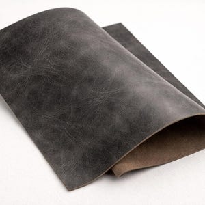 May include: A folded sheet of dark gray leather with a textured surface. The leather has a distressed appearance, with variations in color and tone. The underside of the leather is a lighter brown color.