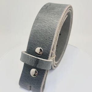 May include: A grey leather belt featuring a textured surface and a smooth, lighter-colored underside. The belt is secured with two polished silver-toned metal snaps on the strap and includes a matching grey leather keeper loop for adjustment.