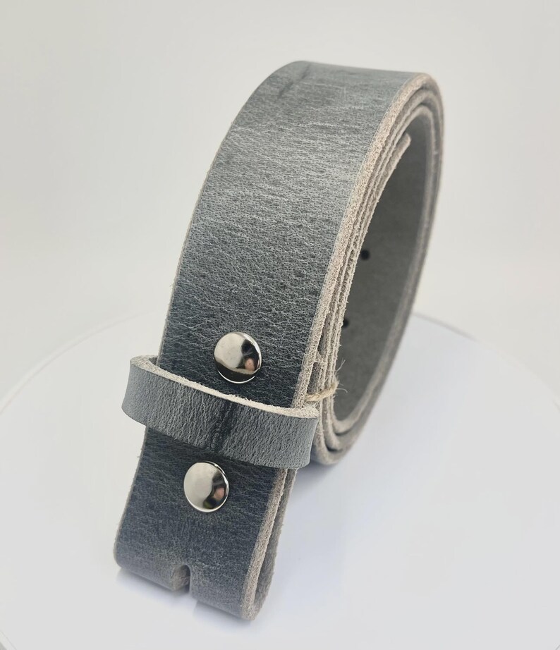 May include: A grey leather belt featuring a textured surface and a smooth, lighter-colored underside. The belt is secured with two polished silver-toned metal snaps on the strap and includes a matching grey leather keeper loop for adjustment.