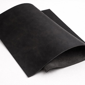 May include: A sheet of black leather, folded over to reveal the lighter underside. The leather has a matte finish and appears to be of high quality. The image is well-lit, highlighting the texture and color of the material.