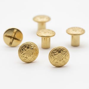 May include: A collection of gold-colored metal rivets and screws. The rivets have a decorative floral pattern on the top. One screw has a cross-shaped indentation. The hardware is displayed on a white background.