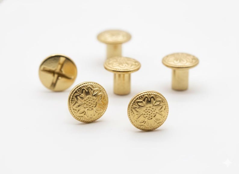 May include: A collection of gold-colored metal rivets and screws. The rivets have a decorative floral pattern on the top. One screw has a cross-shaped indentation. The hardware is displayed on a white background.