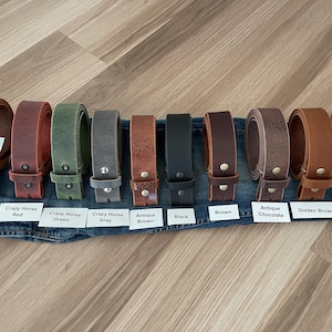Puede incluir: Una colecci&oacute;n de cinturones de cuero en varios colores, incluyendo marr&oacute;n, rojo, verde, gris y negro. Cada cintur&oacute;n se muestra con una hebilla met&aacute;lica y una etiqueta que indica su nombre de color. Los cinturones est&aacute;n colocados sobre una superficie de mezclilla.