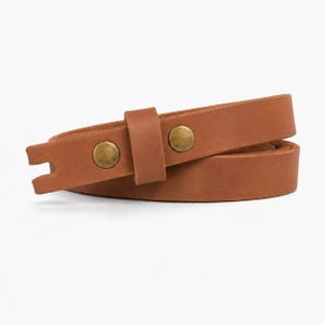 May include: A tan leather strap coiled into a loop, featuring a smooth matte finish and two circular brass-toned metal studs. The strap includes a matching leather keeper loop and a notched end, showcasing a simple and functional design.