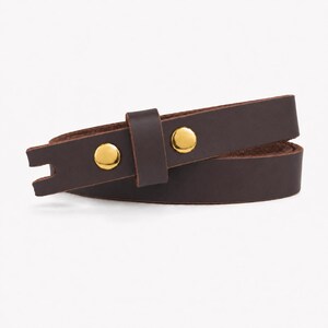 May include: A dark brown leather strap featuring two polished gold-tone metal snap buttons and a matching leather keeper loop. The strap has a notched end and is coiled into a circular shape, showcasing the smooth texture of the leather material.