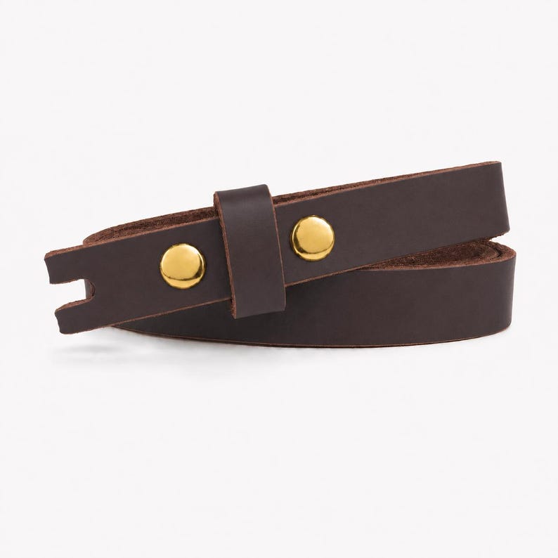 May include: A dark brown leather strap featuring two polished gold-tone metal snap buttons and a matching leather keeper loop. The strap has a notched end and is coiled into a circular shape, showcasing the smooth texture of the leather material.