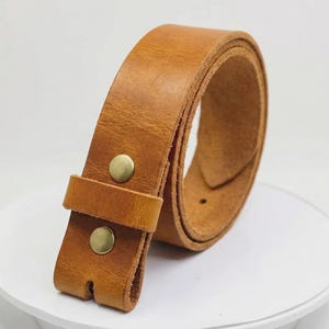 May include: A tan leather belt featuring a smooth, matte finish and a single leather keeper loop. The belt is secured with two circular, metallic brass-colored snap buttons positioned vertically on the front strap section.