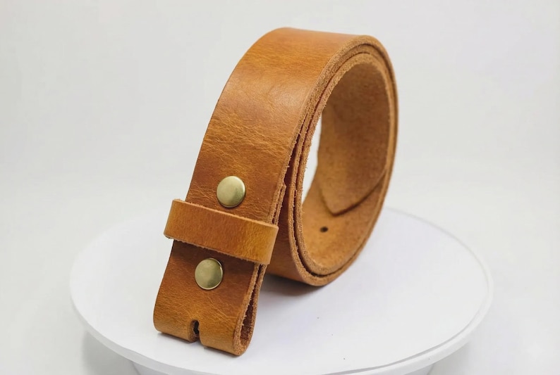 May include: A tan leather belt featuring a smooth, matte finish and a single leather keeper loop. The belt is secured with two circular, metallic brass-colored snap buttons positioned vertically on the front strap section.