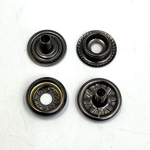 May include: Four metal snap fasteners in a gunmetal finish are arranged on a white surface. Two are open, showing the inner workings, while the other two are closed. One has the text "HATO" around the center.