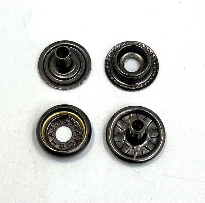 May include: Four metal snap fasteners in a gunmetal finish are arranged on a white surface. Two are open, showing the inner workings, while the other two are closed. One has the text "HATO" around the center.