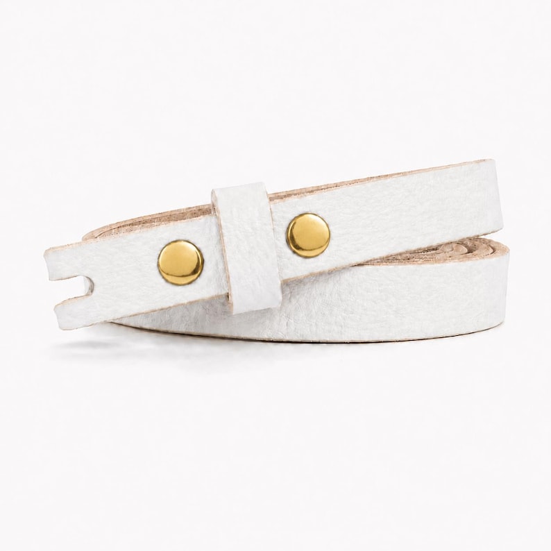May include: A white leather wrap bracelet featuring a textured surface and two polished gold-tone metal studs. The strap is coiled in a circular shape, showing a smooth tan underside and a notched end piece designed for a secure closure.