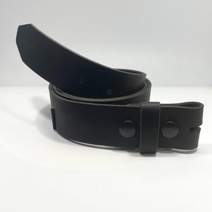 May include: A solid black leather belt coiled in a circular shape. The belt features a smooth matte finish, a pointed tip, and two dark metal snaps near the end to secure the strap. The design includes a single matching black leather keeper loop.