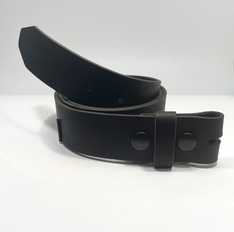 May include: A solid black leather belt coiled in a circular shape. The belt features a smooth matte finish, a pointed tip, and two dark metal snaps near the end to secure the strap. The design includes a single matching black leather keeper loop.