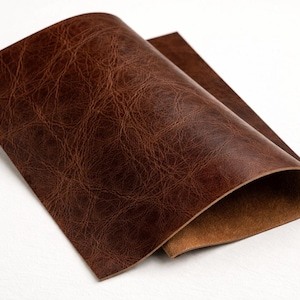 May include: A sheet of brown leather with a textured, distressed appearance. The leather is folded over, revealing a lighter brown underside. The material has a rich, warm tone, suggesting it could be used for crafting.
