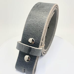 Op de afbeelding: Een grijze leren riem met een zilveren gesp. De riem is opgerold, wat de textuur van het leer en de zilveren klinknagels laat zien. De riem heeft een neutrale kleur, geschikt voor diverse outfits.