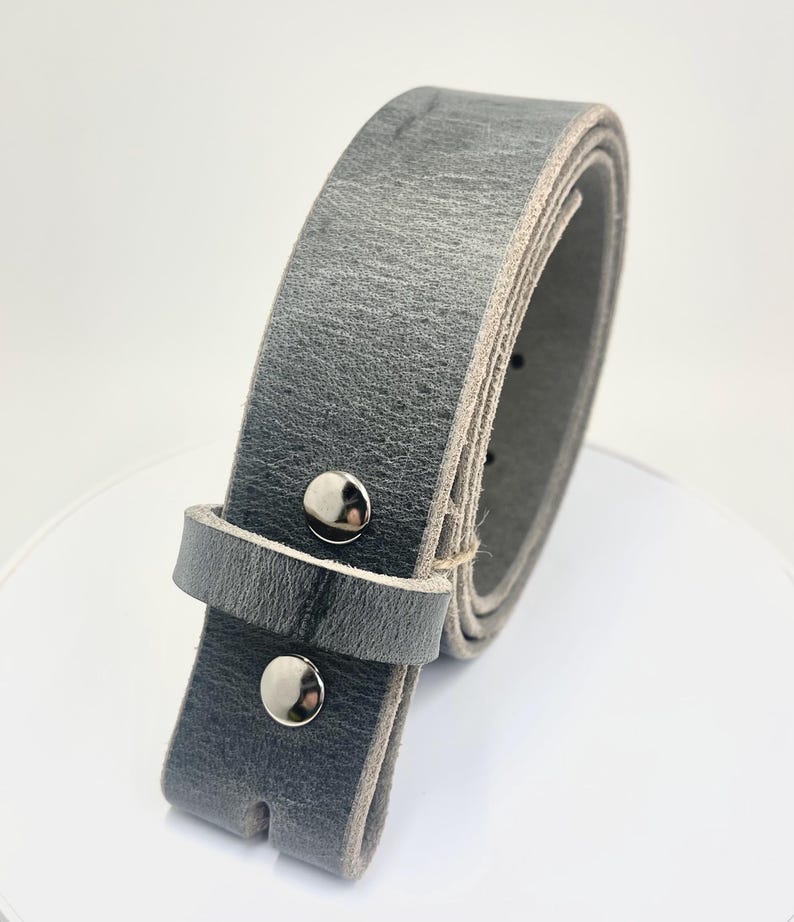 Op de afbeelding: Een grijze leren riem met een zilveren gesp. De riem is opgerold, wat de textuur van het leer en de zilveren klinknagels laat zien. De riem heeft een neutrale kleur, geschikt voor diverse outfits.