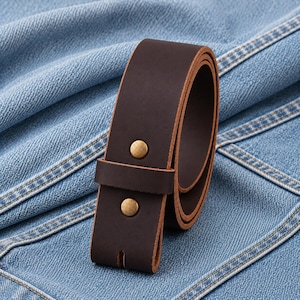 May include: A rolled-up, dark brown leather belt with two brass-colored rivets. The belt is unbuckled and ready for a buckle to be attached. The background is a light blue denim fabric.