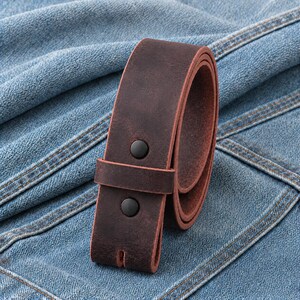 May include: A rolled, dark brown leather belt with two black snap closures. The belt is made of a distressed leather material and is set against a blue denim background.